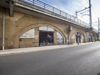 the empty street is next to a brick building and a bridge that has graffiti on it