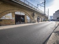 the empty street is next to a brick building and a bridge that has graffiti on it