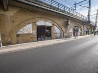 the empty street is next to a brick building and a bridge that has graffiti on it