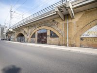 the empty street is next to a brick building and a bridge that has graffiti on it