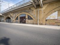 the empty street is next to a brick building and a bridge that has graffiti on it