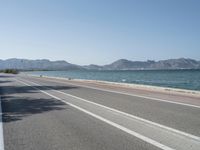 an empty street next to the ocean and mountains with a person walking on the road