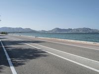 an empty street next to the ocean and mountains with a person walking on the road