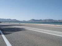 an empty street next to the ocean and mountains with a person walking on the road