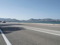 an empty street next to the ocean and mountains with a person walking on the road