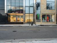 an empty street with store fronts along side a sidewalk that is partly shaded in by glass