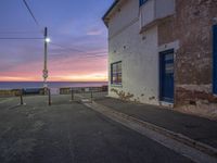 an empty street with an ocean view at sunset near the building with a bench and telephone line