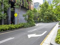 an empty street with a yellow arrow sign near tall buildings and lush green trees lining the street