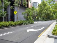 an empty street with a yellow arrow sign near tall buildings and lush green trees lining the street