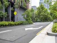 an empty street with a yellow arrow sign near tall buildings and lush green trees lining the street