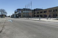 an empty street in front of a commercial building with flags flying from the ground and two trees at the corner