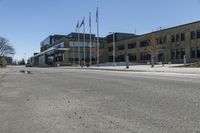 an empty street in front of a commercial building with flags flying from the ground and two trees at the corner