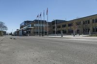 an empty street in front of a commercial building with flags flying from the ground and two trees at the corner