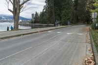 an empty street next to a body of water near some trees and a sidewalk with people walking