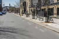 the view of an empty street from across the street on a sunny day with buildings, cars and bicycle racks