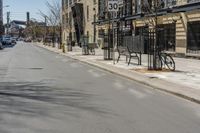 the view of an empty street from across the street on a sunny day with buildings, cars and bicycle racks