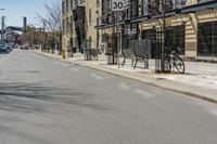 the view of an empty street from across the street on a sunny day with buildings, cars and bicycle racks