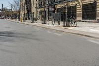 the view of an empty street from across the street on a sunny day with buildings, cars and bicycle racks