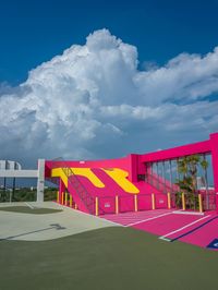 an empty tennis court with pink and yellow on it, under a blue sky with clouds