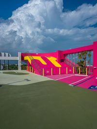 an empty tennis court with pink and yellow on it, under a blue sky with clouds