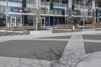 an empty area near buildings is shown with trees in the foreground, and several pedestrians walk on the pavement