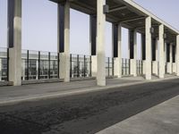 an empty walkway near the parking lot with concrete columns and glass doors, next to a large building with many windows