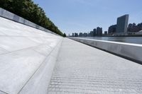 an empty walkway at the waterfront next to water and skyscrapers in the background of a sunny day