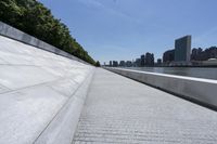 an empty walkway at the waterfront next to water and skyscrapers in the background of a sunny day