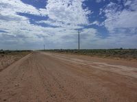 a dirt road with clouds and telephone poles in the distance in the desert with grass