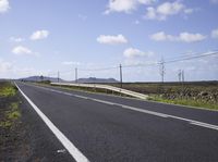 a empty highway running between a field with power lines above it and a rock wall with two people on either side