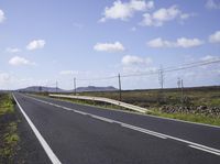 a empty highway running between a field with power lines above it and a rock wall with two people on either side