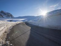 a road in the mountains in the sun covered by snow with a snow plow on it