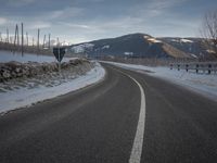 an empty road with snow covering mountains and trees behind it on a cloudy day in winter