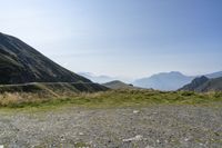 a rocky mountain with green grass, with mountains in the background and a few small hills