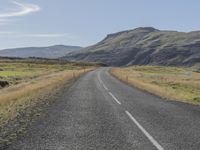 an empty road with a grassy field beside it and a fence to the side of the road, with mountains in the background