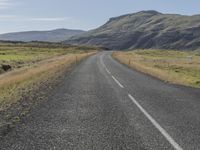 an empty road with a grassy field beside it and a fence to the side of the road, with mountains in the background