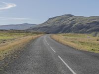 an empty road with a grassy field beside it and a fence to the side of the road, with mountains in the background