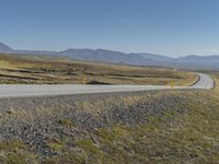 a paved road stretches into the distance through a valley filled with mountains and grass, with a lone yellow sign that reads stop to wait