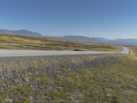 a paved road stretches into the distance through a valley filled with mountains and grass, with a lone yellow sign that reads stop to wait
