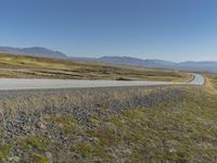 a paved road stretches into the distance through a valley filled with mountains and grass, with a lone yellow sign that reads stop to wait