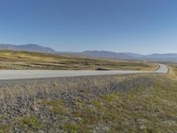 a paved road stretches into the distance through a valley filled with mountains and grass, with a lone yellow sign that reads stop to wait