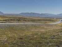 a paved road stretches into the distance through a valley filled with mountains and grass, with a lone yellow sign that reads stop to wait