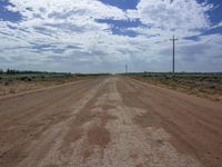 the open dirt road in a deserted landscape under a sky filled with white clouds and telephone poles