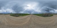 a motorcycle track with gravel and clouds in the sky behind it a green field with bushes in front of it