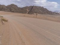 an empty dirt road between large mountain range and a grassy plain with gravel floor and grass in foreground