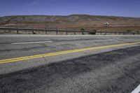 two yellow lines are on the road beside a fence and mountains in the distance of the photo