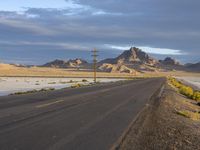 a desert road with some mountains in the background, with a few telephone lines above and yellow grass on each side