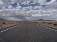 an empty highway in the middle of nowhere with mountains in the background and a cloudy sky overhead