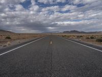 an empty highway in the middle of nowhere with mountains in the background and a cloudy sky overhead