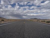 an empty highway in the middle of nowhere with mountains in the background and a cloudy sky overhead